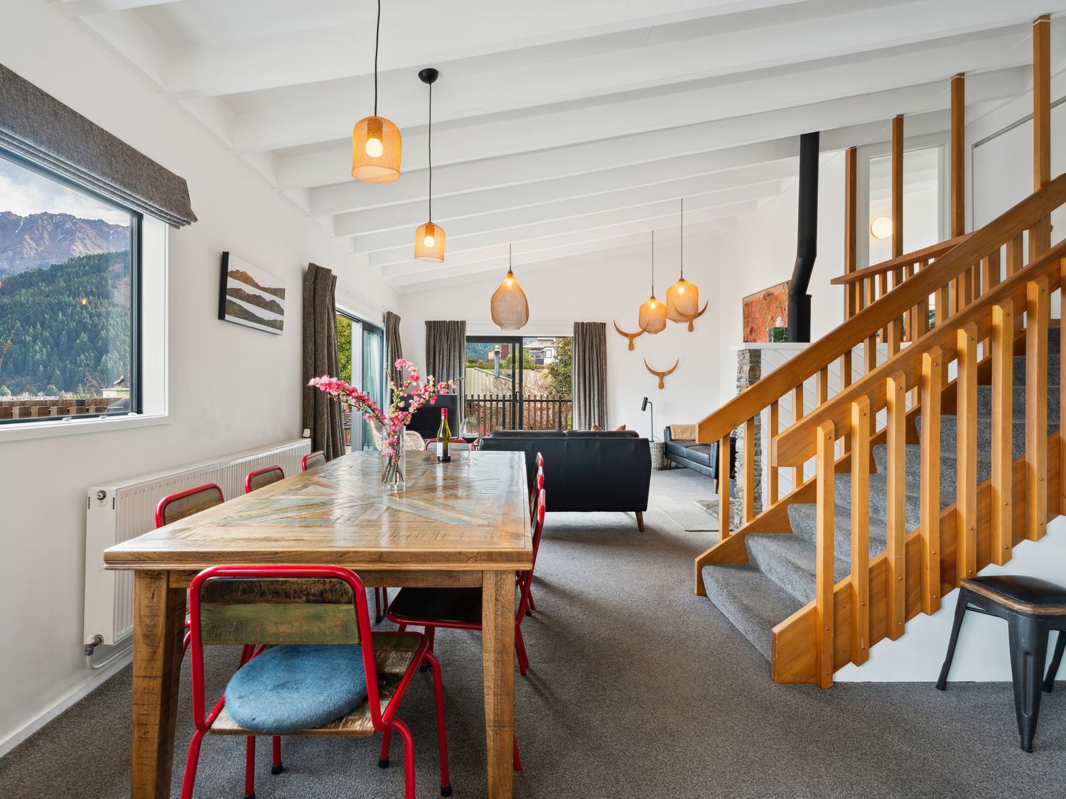 A dining and living room with wooden table red chairs pendant lights staircase and mountain view at Central Southern Lakes Queenstown Holiday Home Queenstown