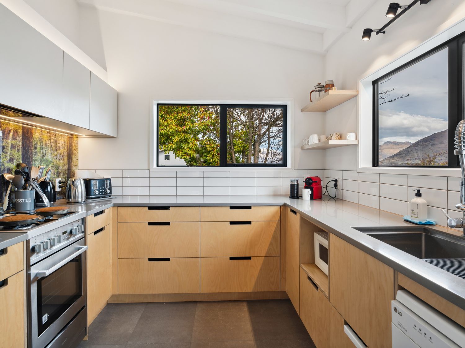 A kitchen with wooden cabinets stainless steel appliances and two windows showing trees and mountains at Central Southern Lakes - Queenstown Holiday Home in Queenstown