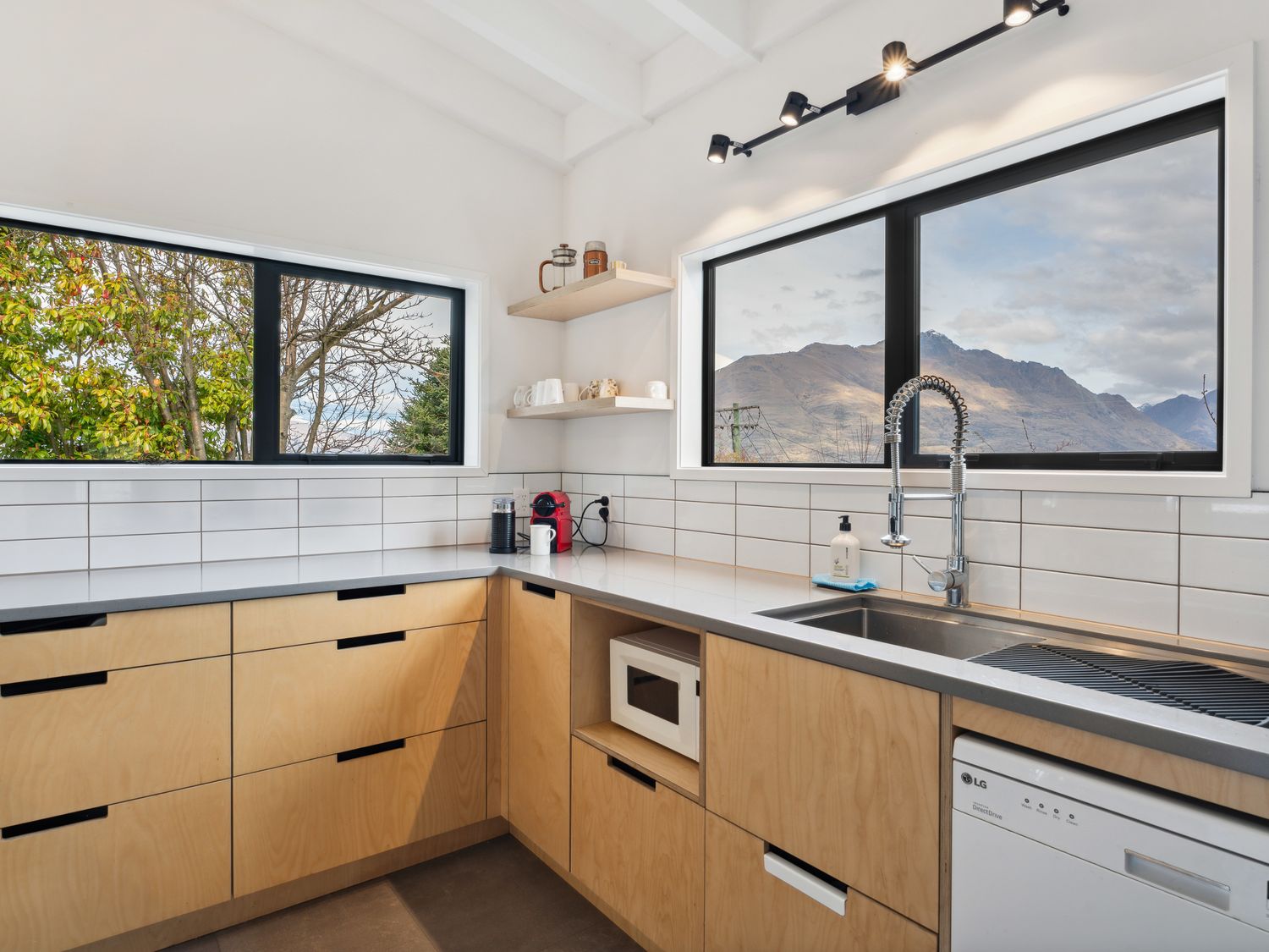 A kitchen with wooden cabinets a microwave and a dishwasher under the counter and windows showing trees and mountains at Central Southern Lakes - Queenstown Holiday Home in Queenstown