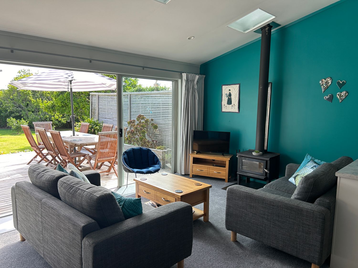 A living room with gray sofas a wood stove a blue wall and a sliding door leading to a patio with a table and chairs at Coro Beach Haven  Hahei Holiday Home