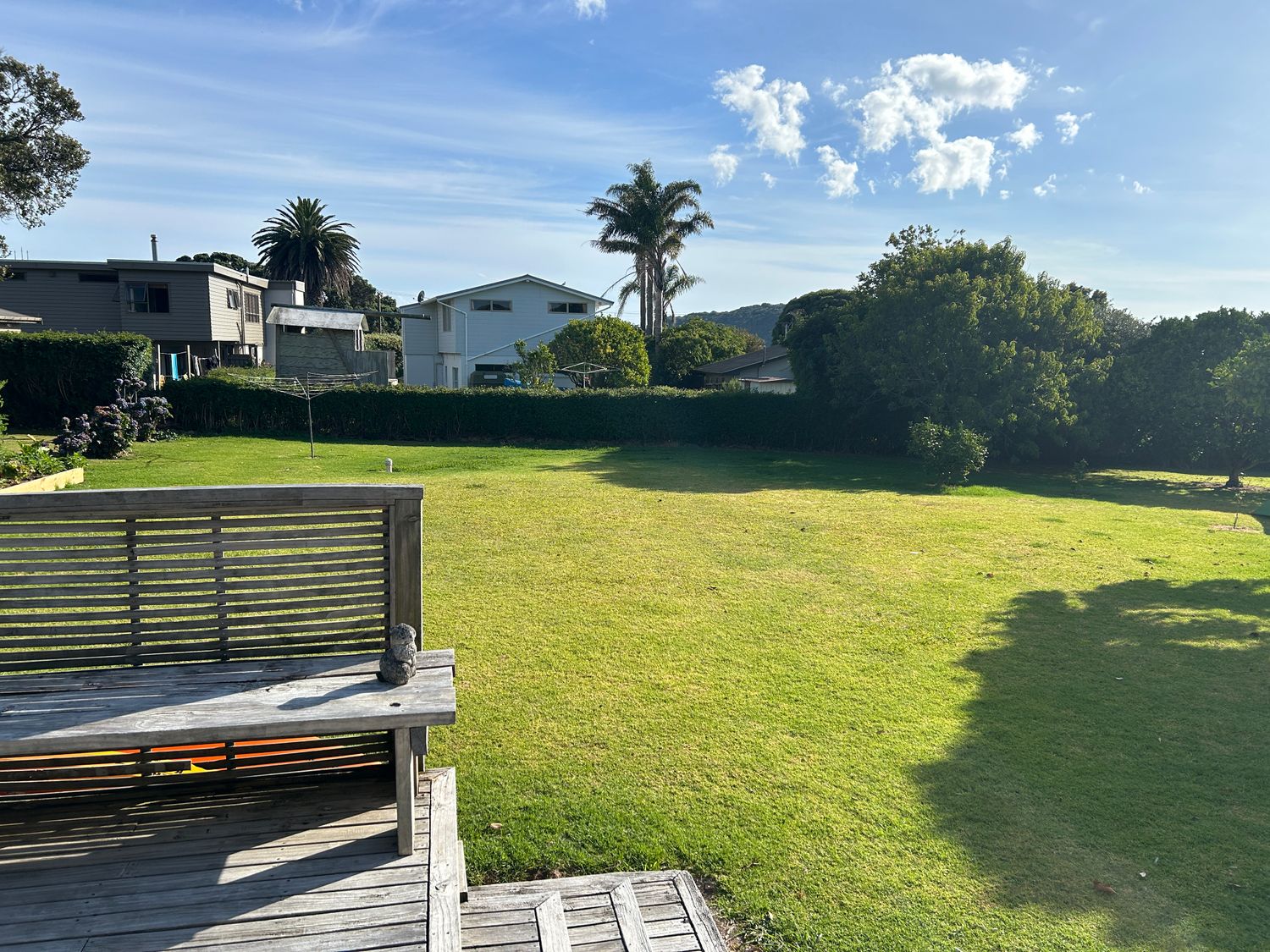 A garden with a wooden bench on a deck and houses and trees in the background at Coro Beach Haven Hahei Holiday Home in Hahei