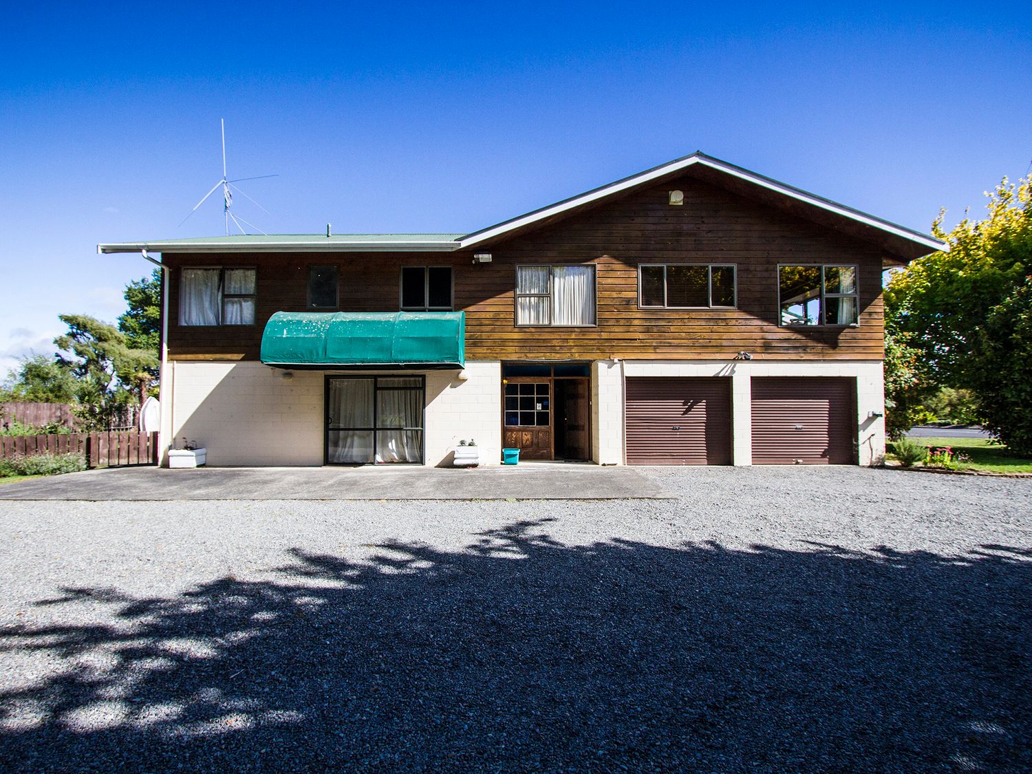 A two-story house with a green awning over a door two garage doors and a gravel driveway at Riverbed Lodge - Lake Taupo Home in Tauranga Taupo
