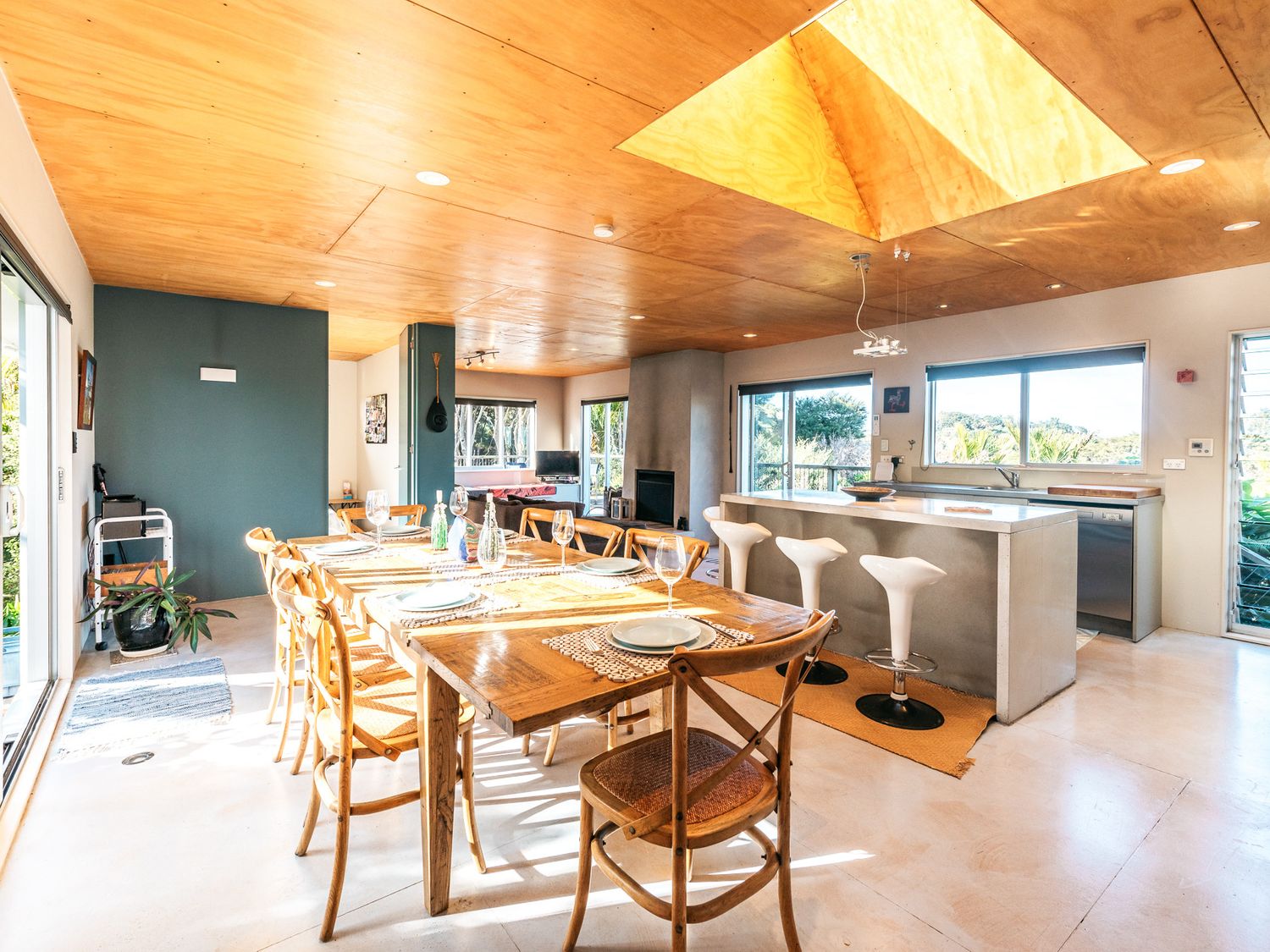 A dining area with a wooden table set with plates and glasses next to a kitchen island with three white bar stools at The Fat Pigeon - Omiha Holiday Home in Rocky Bay