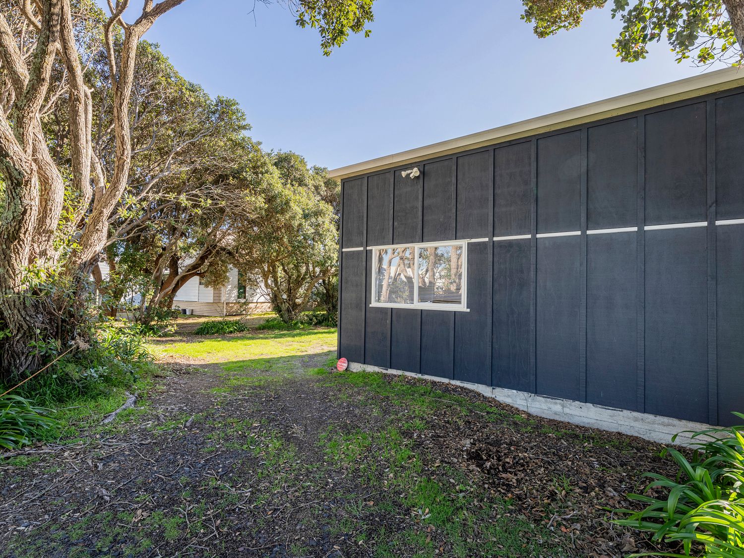 The side of a dark gray building with a window and trees surrounding a dirt and grass pathway at Rangimarie on Kenwood Matarangi Holiday Home in Matarangi