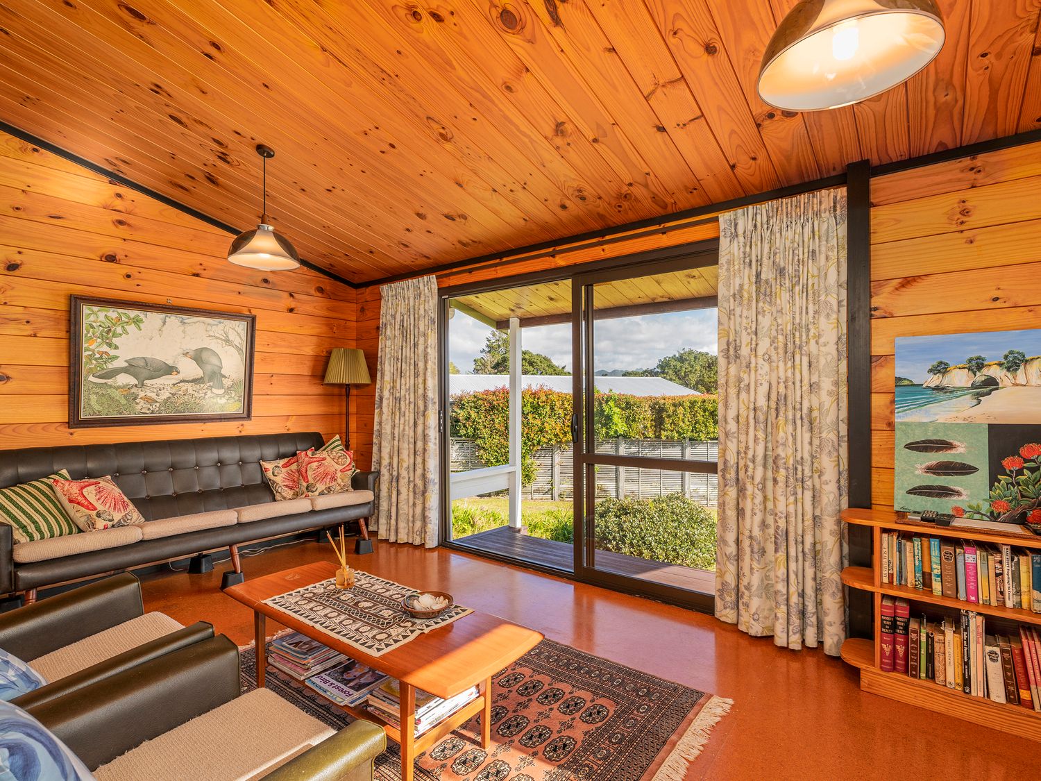 A living room with wooden walls and ceiling a sofa armchairs coffee table bookshelf and sliding glass doors at Retro Revival - Pauanui Holiday Home in Pauanui