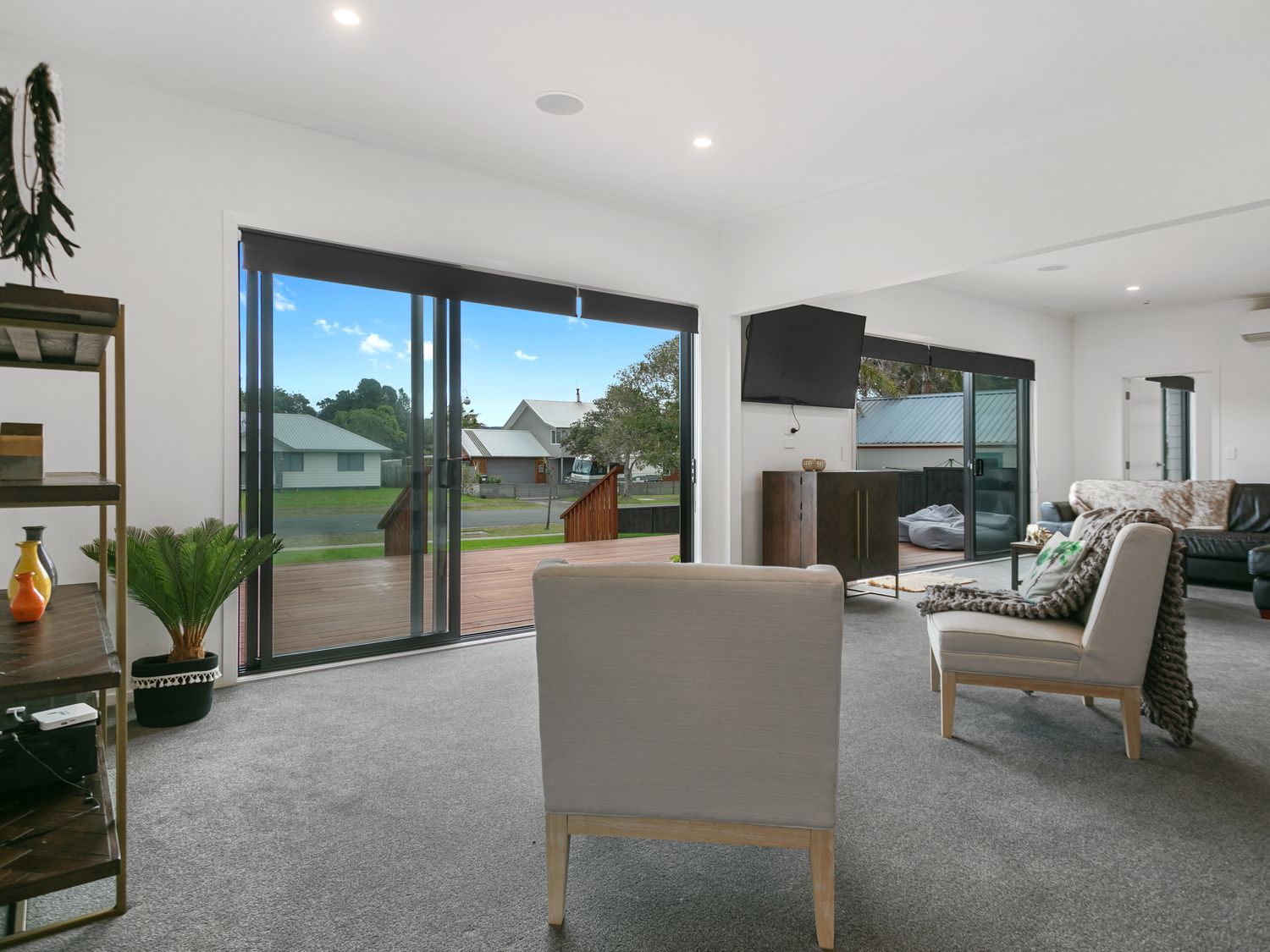 A living room with sliding glass doors a television a light-colored armchair a dark cabinet and a black leather sofa at Sky Above Beach Below - Waihi Beach Holiday Home in Waihi Beach