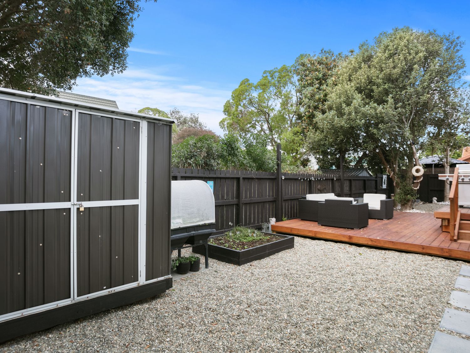 A backyard with a metal shed a raised garden bed outdoor seating on a wooden deck and trees at Sky Above Beach Below - Waihi Beach Holiday Home in Waihi Beach