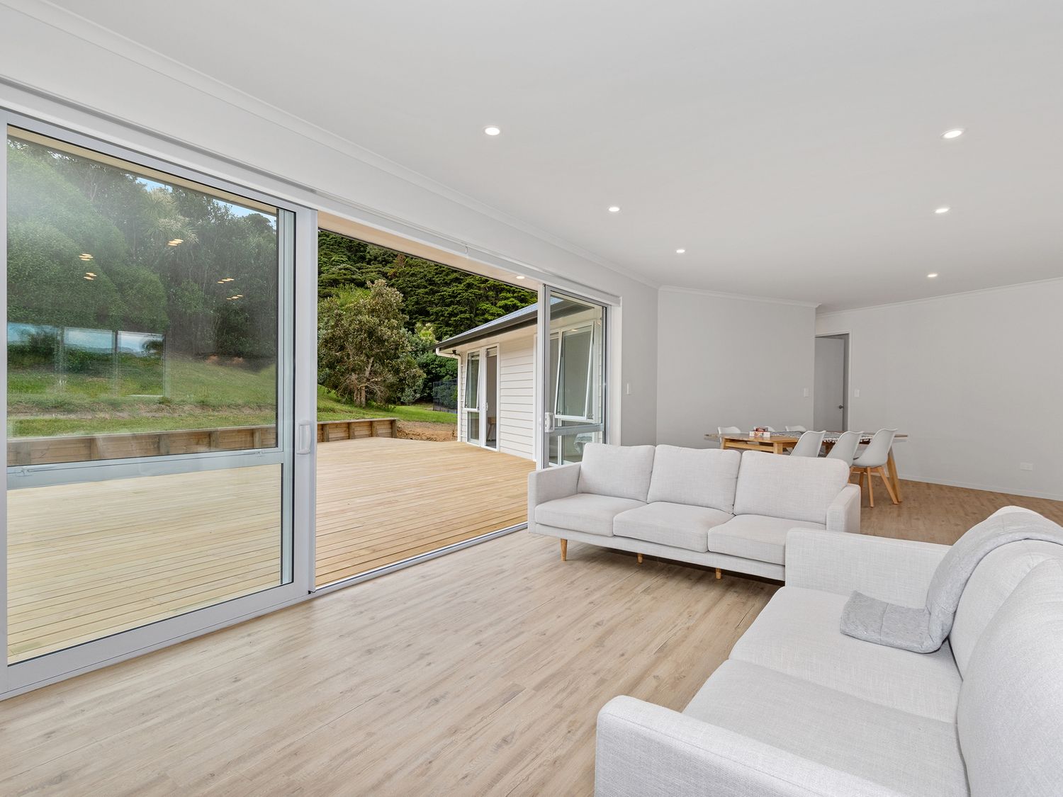 A living room with two white sofas and a dining table with chairs near large sliding glass doors opening to a wooden deck at The Platinum Rendezvous - Matapouri Holiday Home in Matapouri
