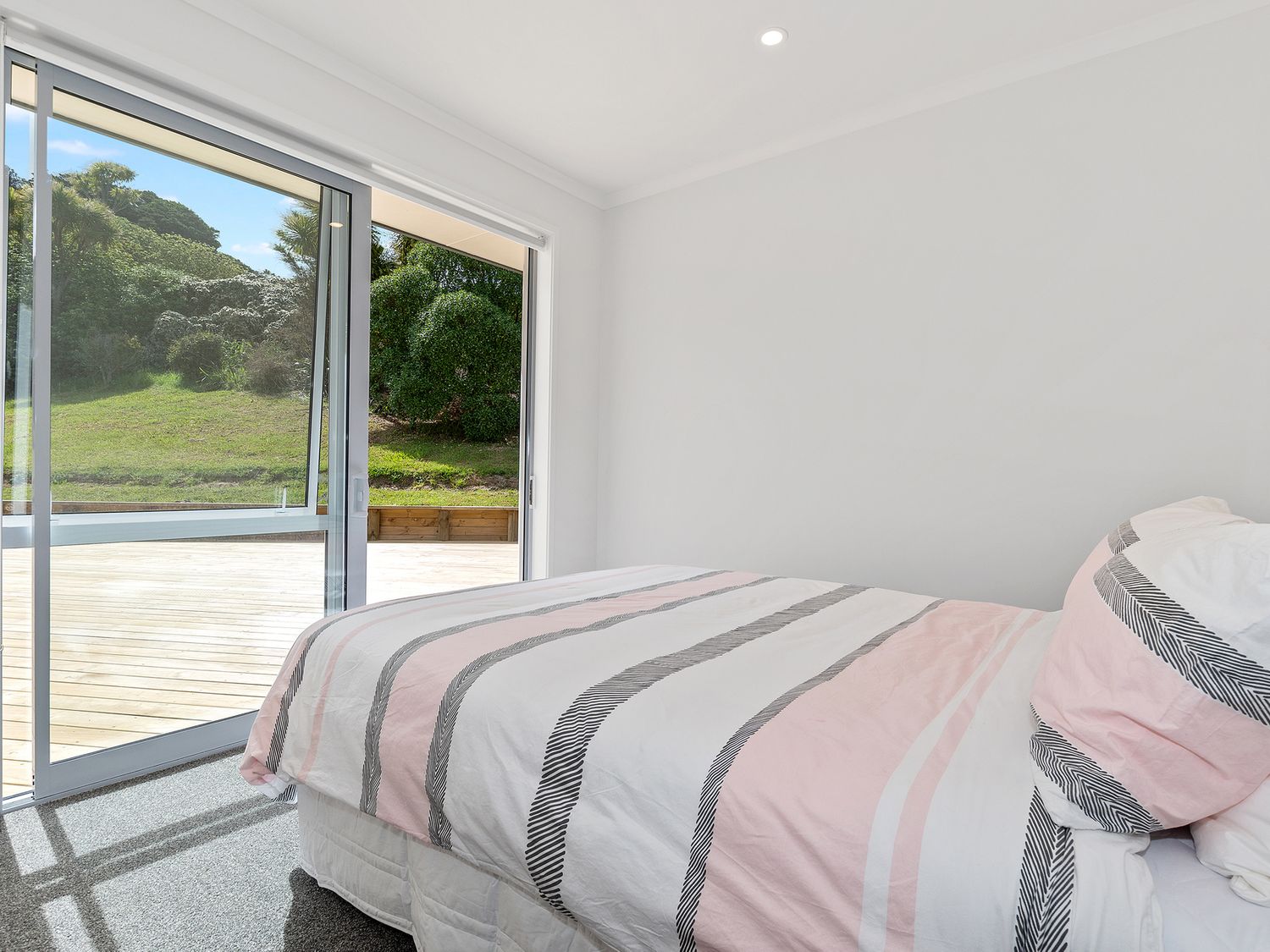 A bedroom with a bed covered in striped bedding next to a sliding glass door showing a wooden deck and greenery outside at The Platinum Rendezvous - Matapouri Holiday Home in Matapouri