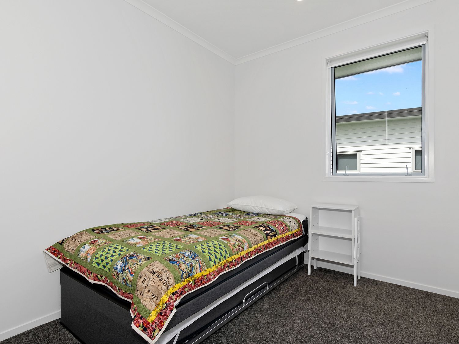 A small bedroom with a single bed covered by a patterned quilt and a small white shelf near a window at The Platinum Rendezvous - Matapouri Holiday Home in Matapouri