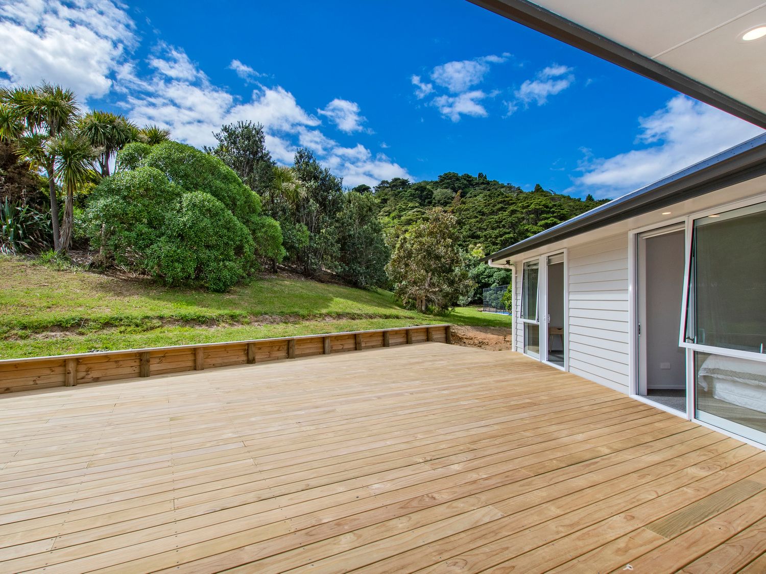 A wooden deck outside a house with sliding glass doors and green trees on a hill at The Platinum Rendezvous - Matapouri Holiday Home in Matapouri