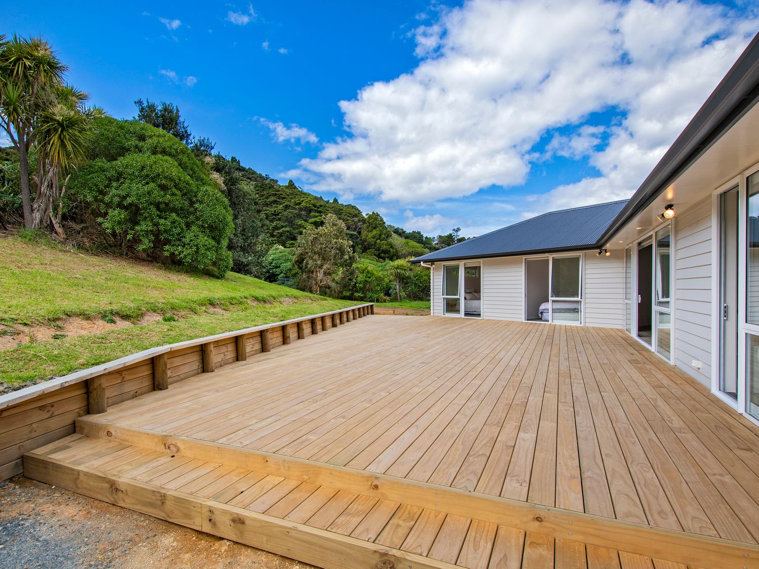 A wooden deck outside a white house with sliding glass doors and grassy hill with trees in the background at The Platinum Rendezvous - Matapouri Holiday Home in Matapouri