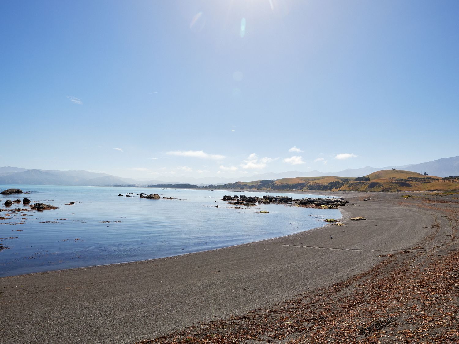 A sandy beach with rocks in the water and hills in the background at The Kaikoura Bach Retreat in Kaikoura