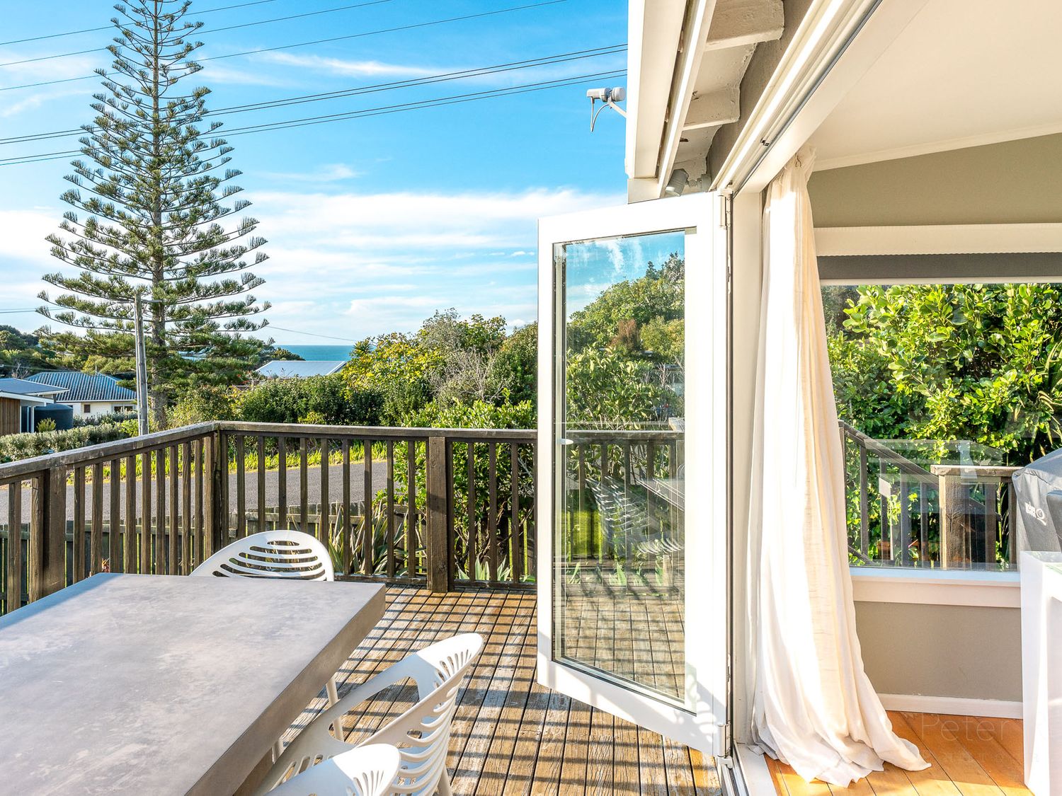 An outdoor wooden deck with a table and chairs next to an open glass door overlooking trees and distant houses at Eden Escape Onetangi