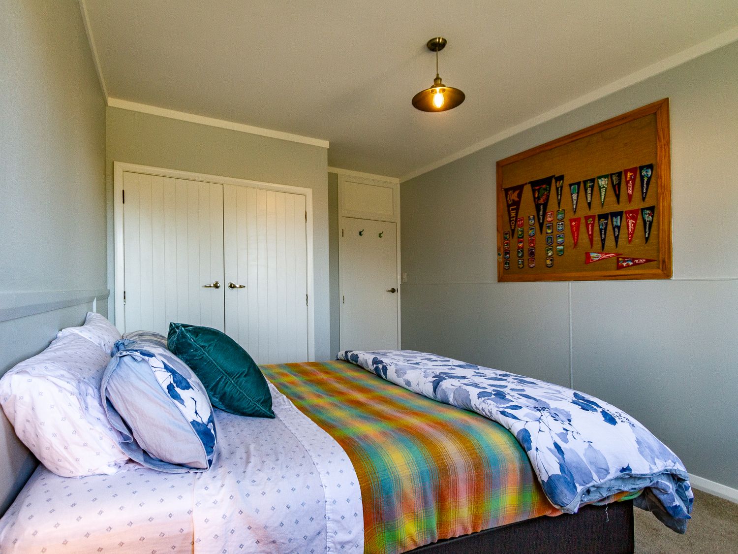 A bedroom with a bed covered with multiple pillows and a colorful blanket a closet with double white doors and a single white door a bulletin board with various flags and badges on the wall Ohakune Barracks - Ohakune Holiday Home Ohakune