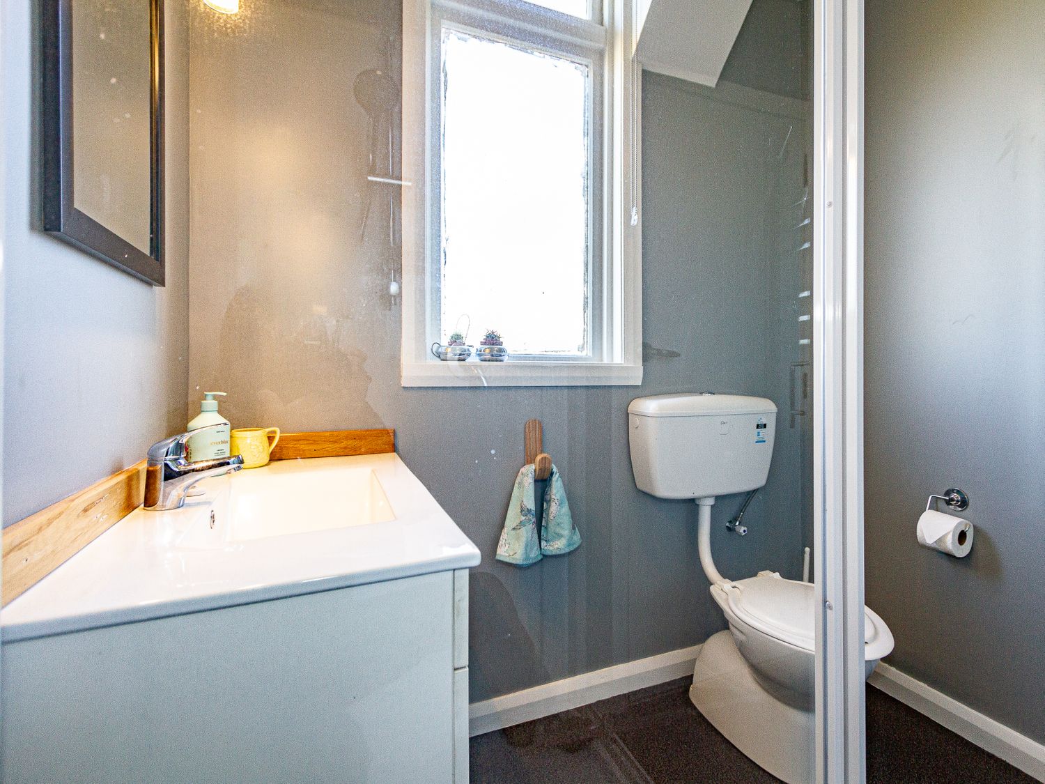 A bathroom with a white sink, mirror, window, toilet, toilet paper holder, and two hanging towels at Ohakune Barracks Ohakune