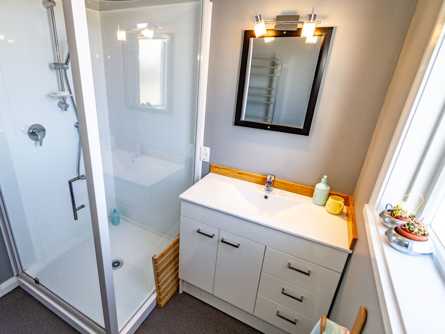 A bathroom with a shower, white sink cabinet, mirror with lights, and small plants on the windowsill at Ohakune Barracks in Ohakune