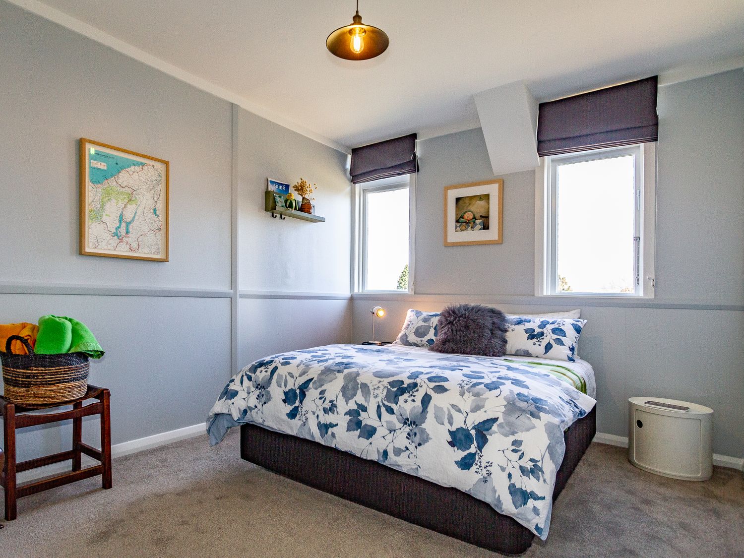A bedroom with a bed with floral bedding between two windows a wooden chair with a basket a map and framed pictures on the walls at Ohakune Barracks in Ohakune