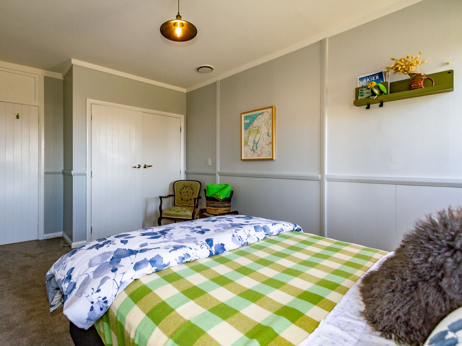 A bedroom with a bed covered in green checkered blanket and floral duvet a chair basket on a small table and wall shelf with decorations at Ohakune Barracks Ohakune
