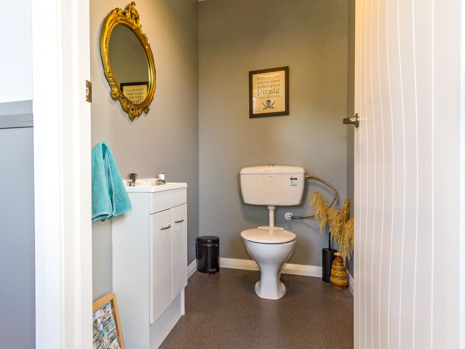 A bathroom with a white toilet and sink cabinet with a turquoise towel hanging an ornate gold mirror on the wall and a framed picture at Ohakune Barracks Ohakune
