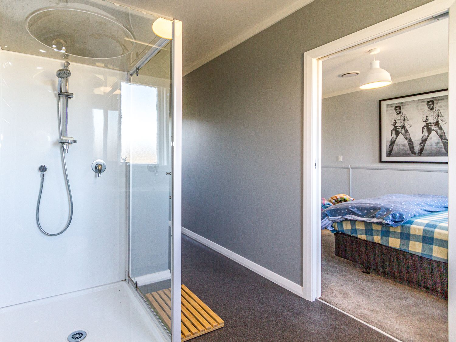A bathroom with a glass shower enclosure next to a hallway leading to a bedroom with a bed and framed artwork at Ohakune Barracks Ohakune