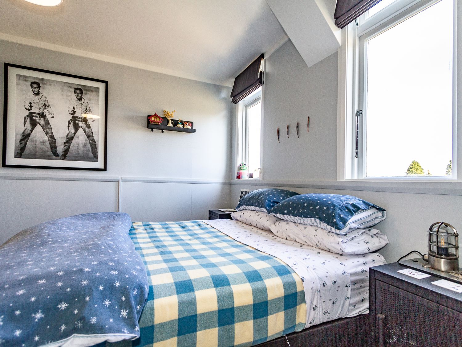 A bedroom with a blue and white check blanket on a bed with pillows next to two windows and a black nightstand with a lantern at Ohakune Barracks Ohakune