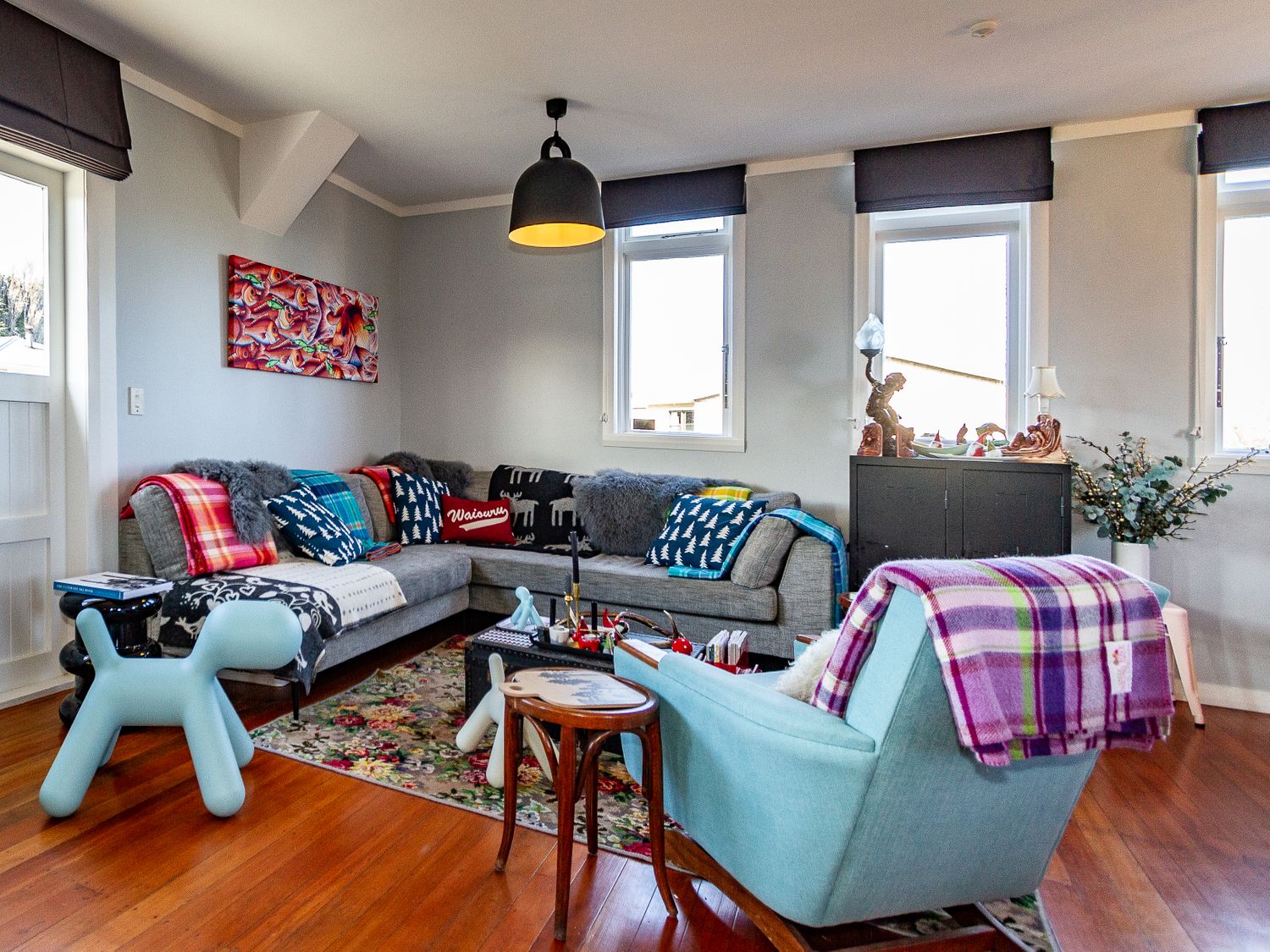 A living room with a grey sectional sofa covered with cushions and blankets a light blue armchair with a purple plaid blanket small wooden side table a decorative blue dog figure a floral rug on wooden floor black cabinet with lamps and decorative items three windows with blinds Ohakune Barracks Ohakune