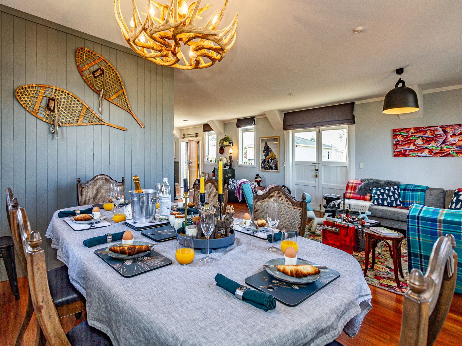 A dining table set with croissants and orange juice in a room with wooden chairs snowshoes on the wall and a living area with sofas and artwork at Ohakune Barracks Ohakune