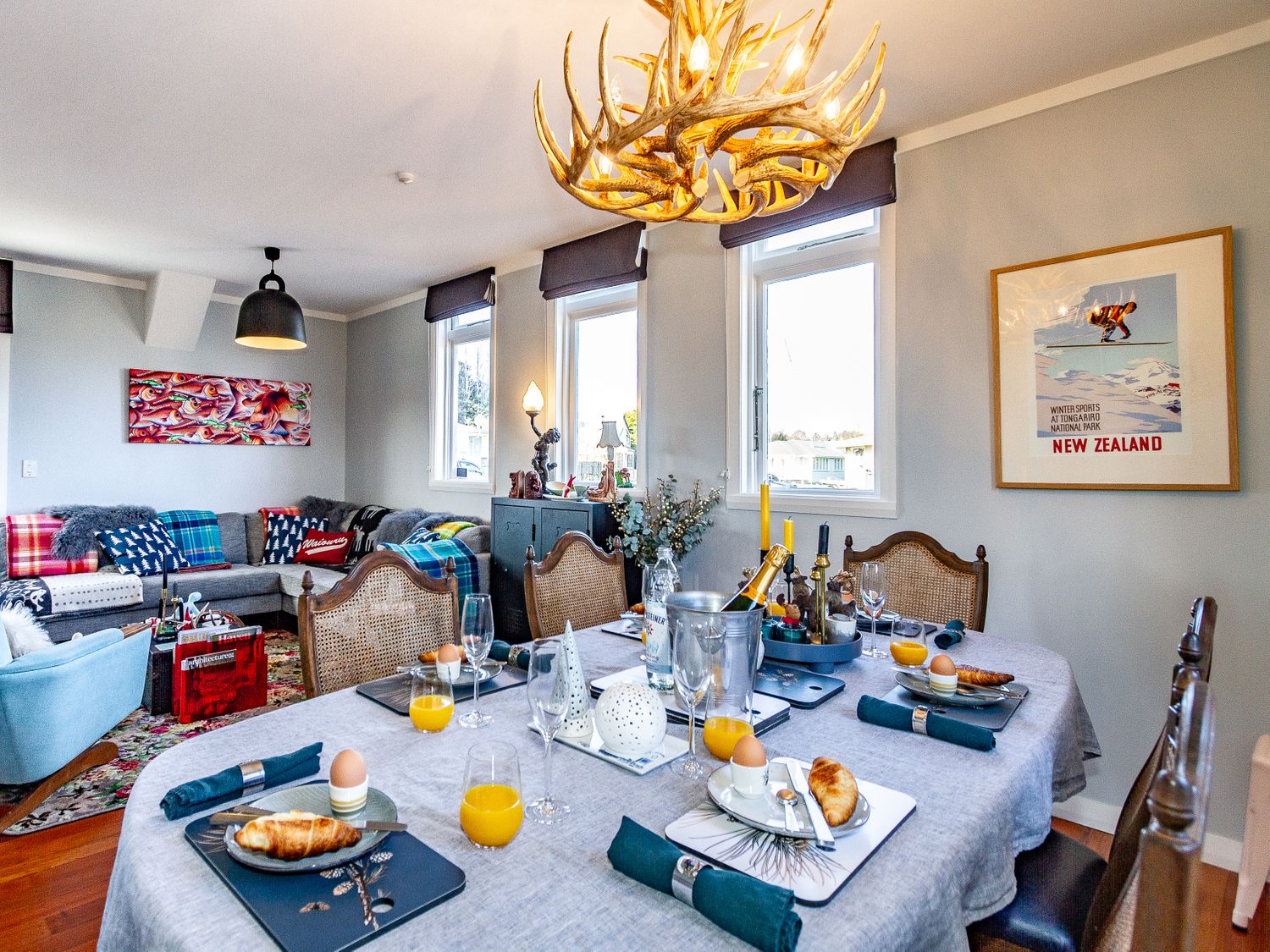 A dining table set with croissants eggs and orange juice in a room with wicker chairs a gray sofa and windows at Ohakune Barracks Ohakune