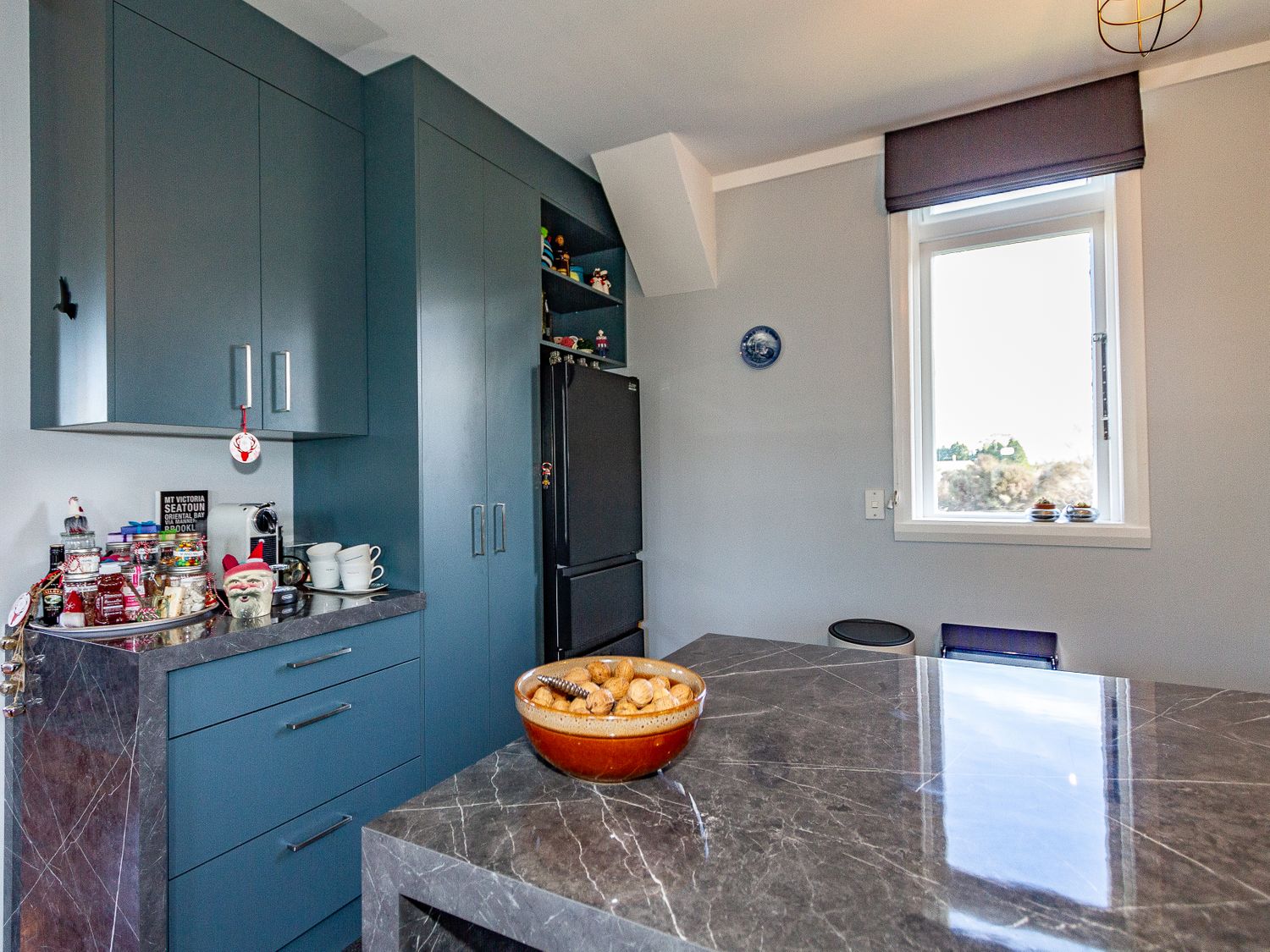A kitchen with blue cabinets a black refrigerator a marble countertop with a bowl of nuts and a window with a dark valance at Ohakune Barracks in Ohakune