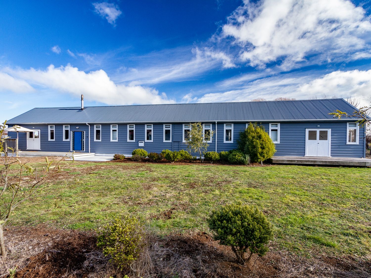 A single story long blue house with multiple windows surrounded by grass and bushes at Ohakune Barracks Ohakune