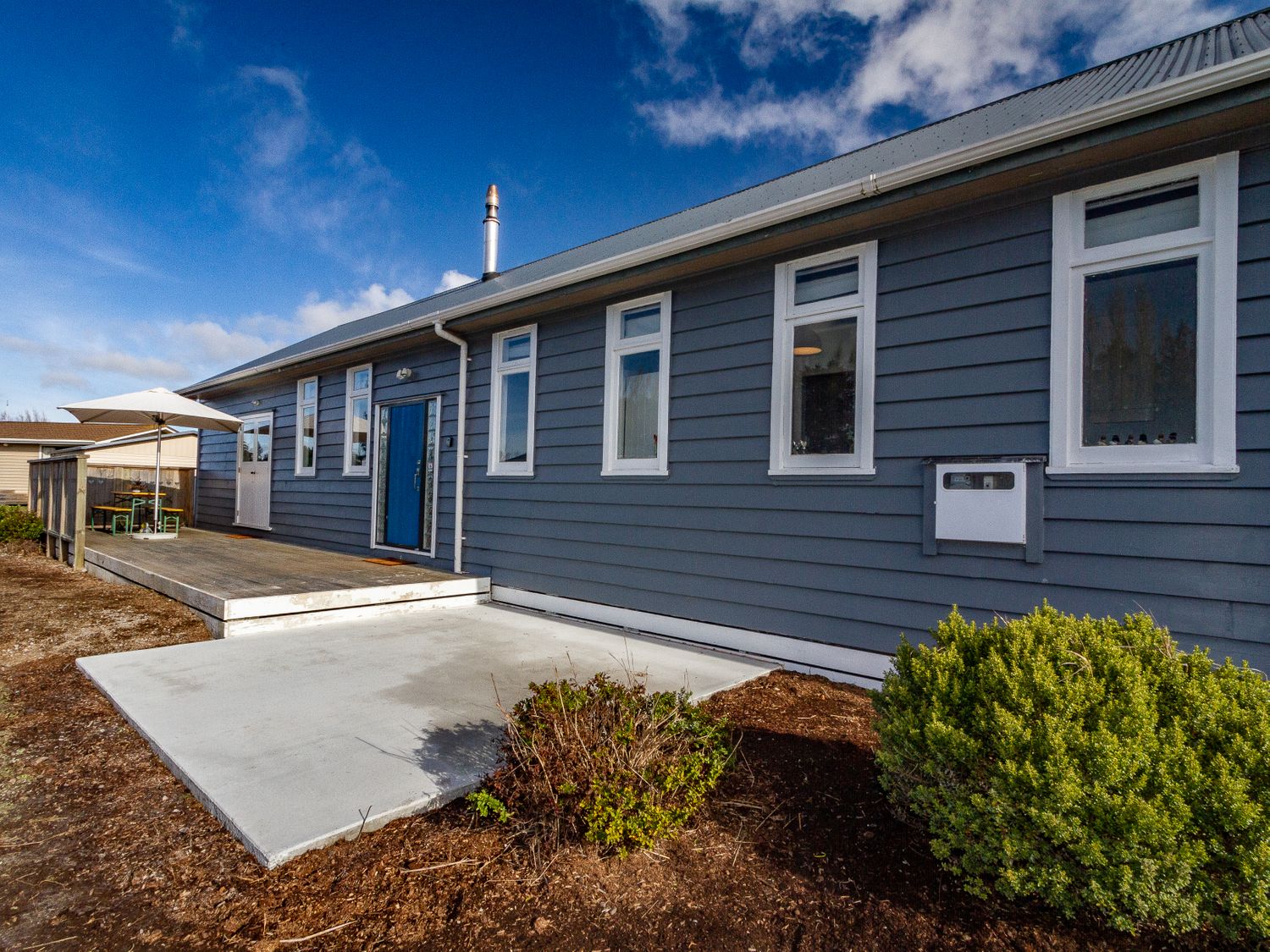 The exterior of a single story house with blue siding multiple windows a blue door a wooden deck with an umbrella and outdoor furniture at Ohakune Barracks Ohakune