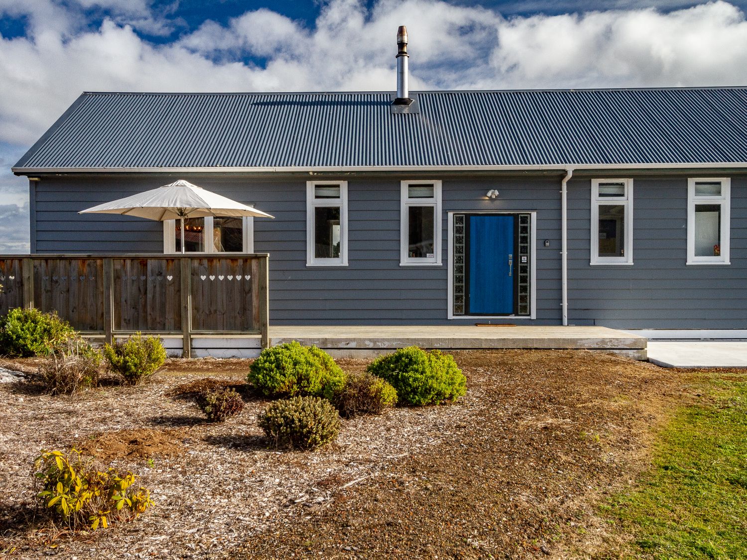 A grey house with a blue front door a wooden fence with a white umbrella and bushes in front at Ohakune Barracks Ohakune