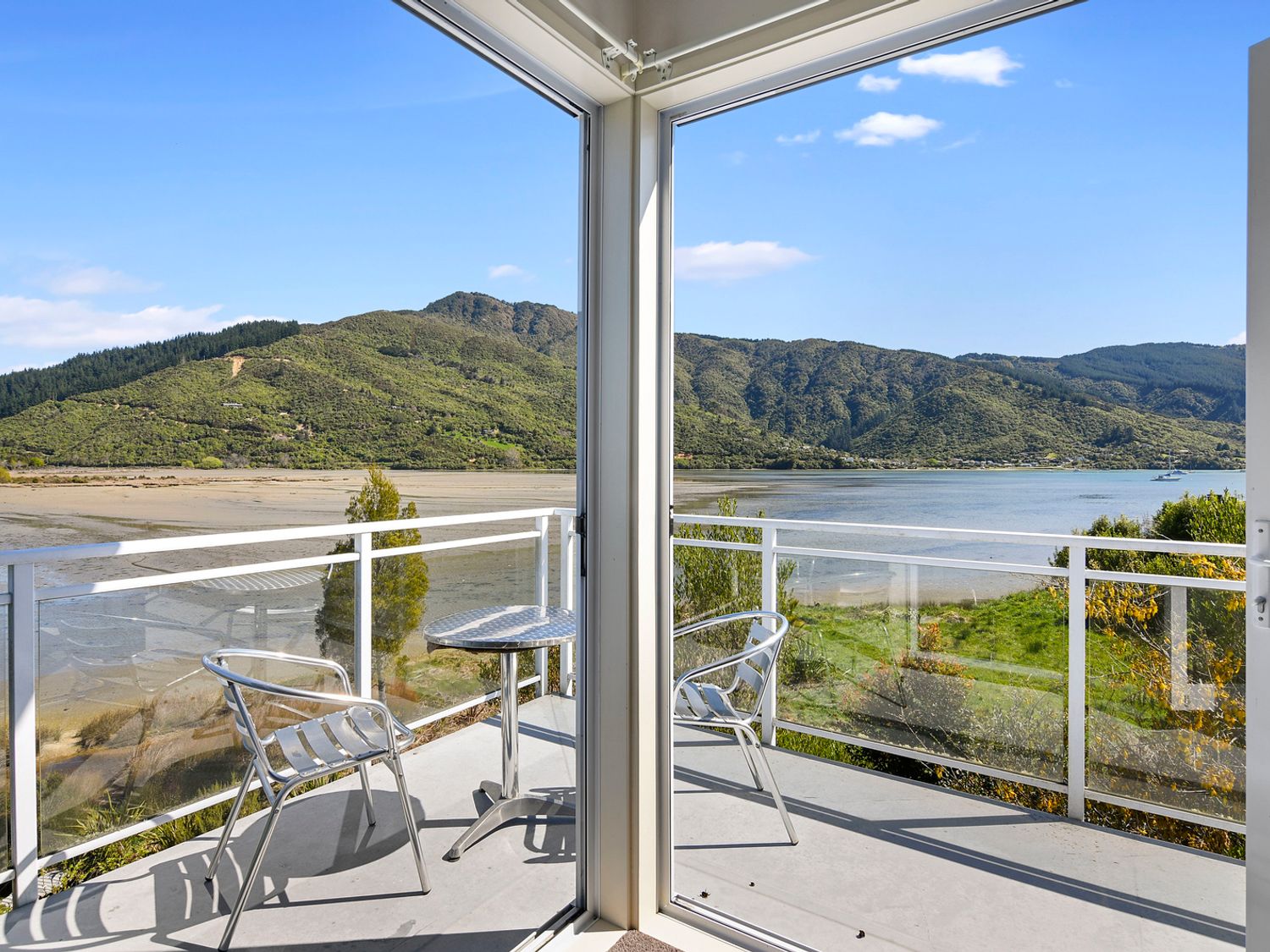 A balcony with two metal chairs and a round metal table overlooking a river and green hills at Millard House - Marlborough Sounds