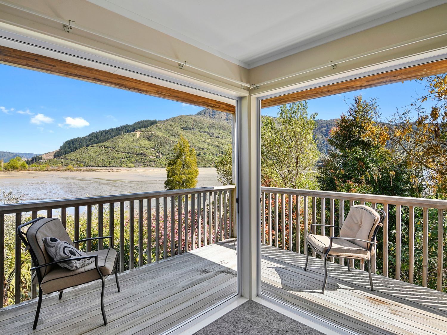 A wooden deck with two cushioned chairs overlooking trees and hills at Millard House - Marlborough Sounds
