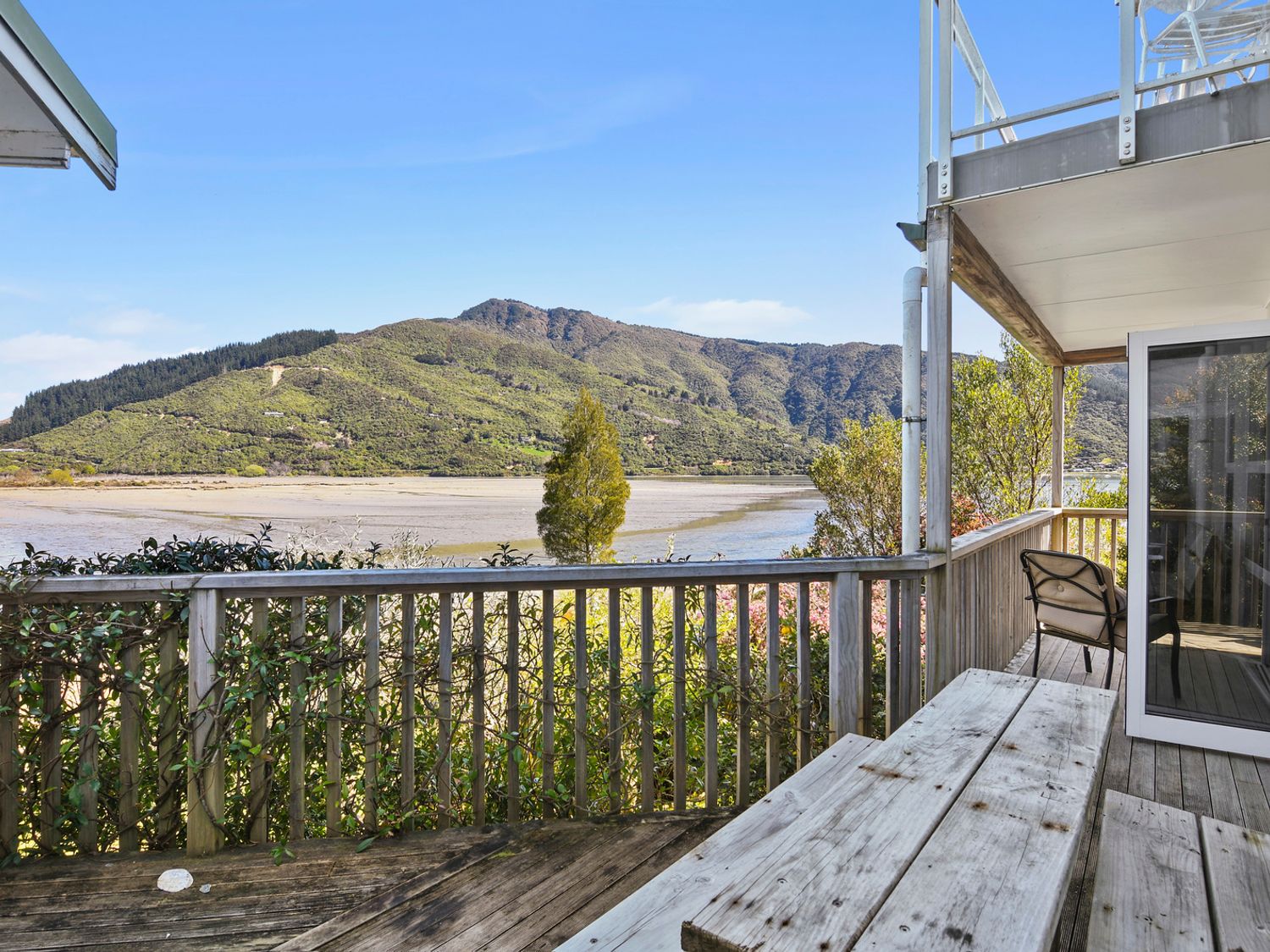 An outdoor wooden deck with picnic table and chair overlooking a river and mountains at Millard House - Marlborough Sounds
