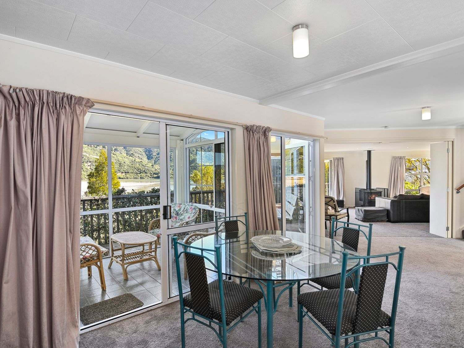 A dining area with a glass table and black chairs next to a sliding glass door leading to a porch with wicker furniture and a view of trees and hills at Millard House - Marlborough Sounds