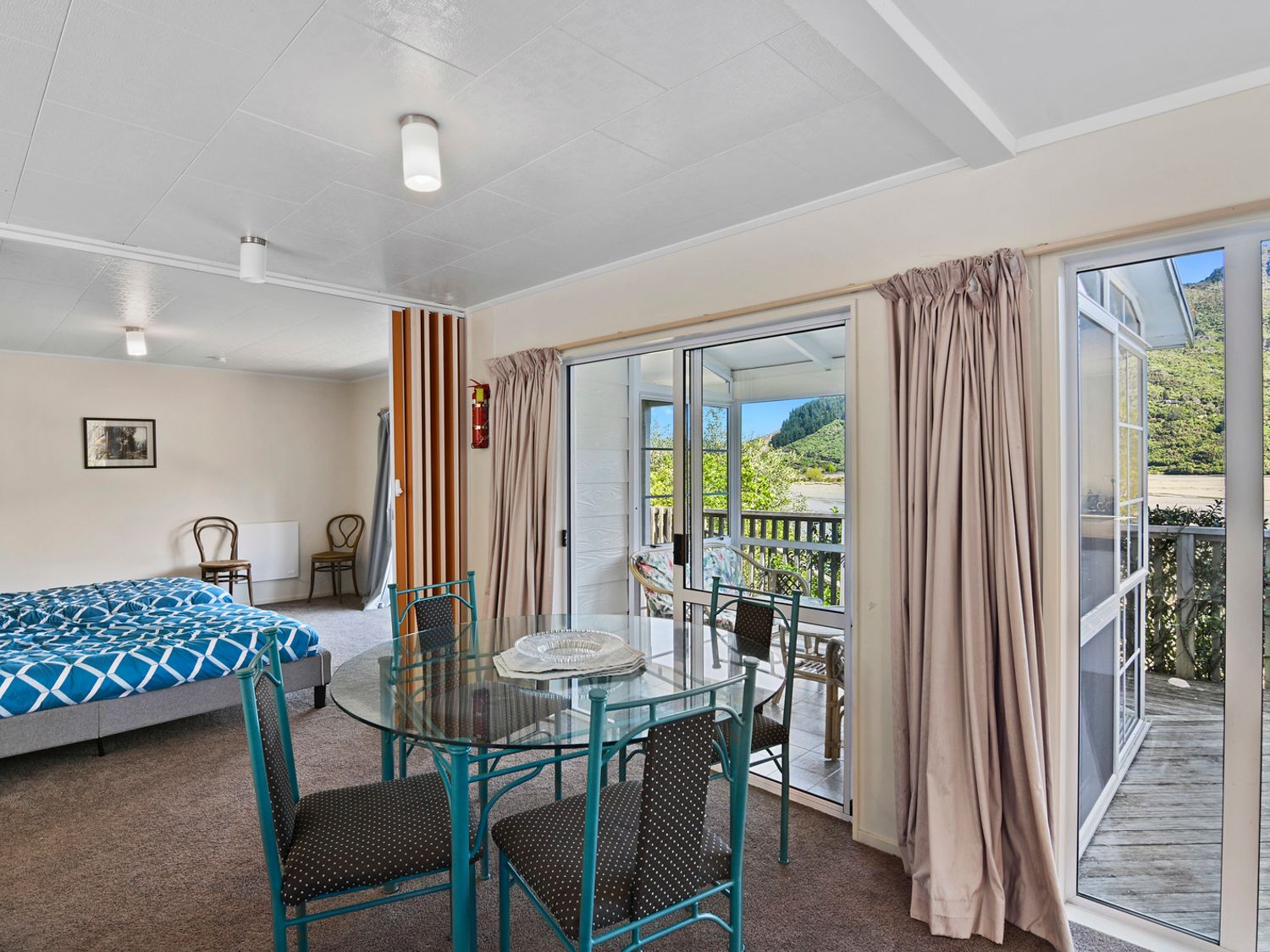 A room with a glass dining table and chairs next to sliding glass doors leading to a balcony with outdoor seating and a bed in the adjacent area at Millard House - Marlborough Sounds