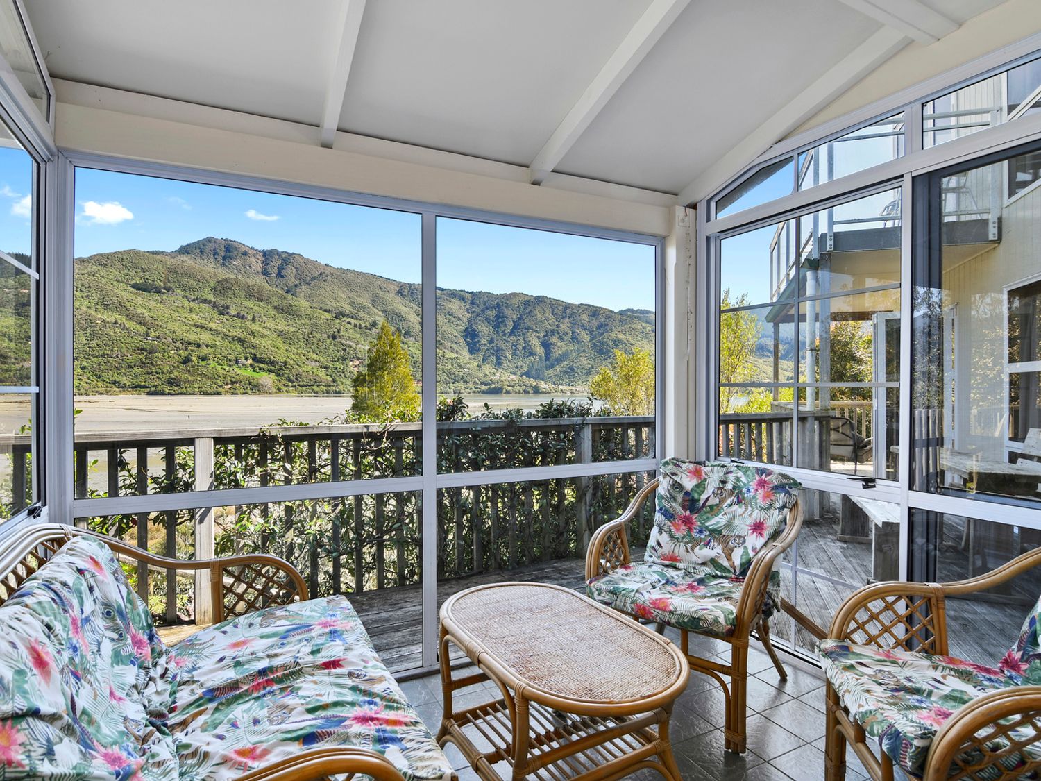 A sunroom with wicker furniture and floral cushions overlooking a mountain landscape at Millard House - Marlborough Sounds