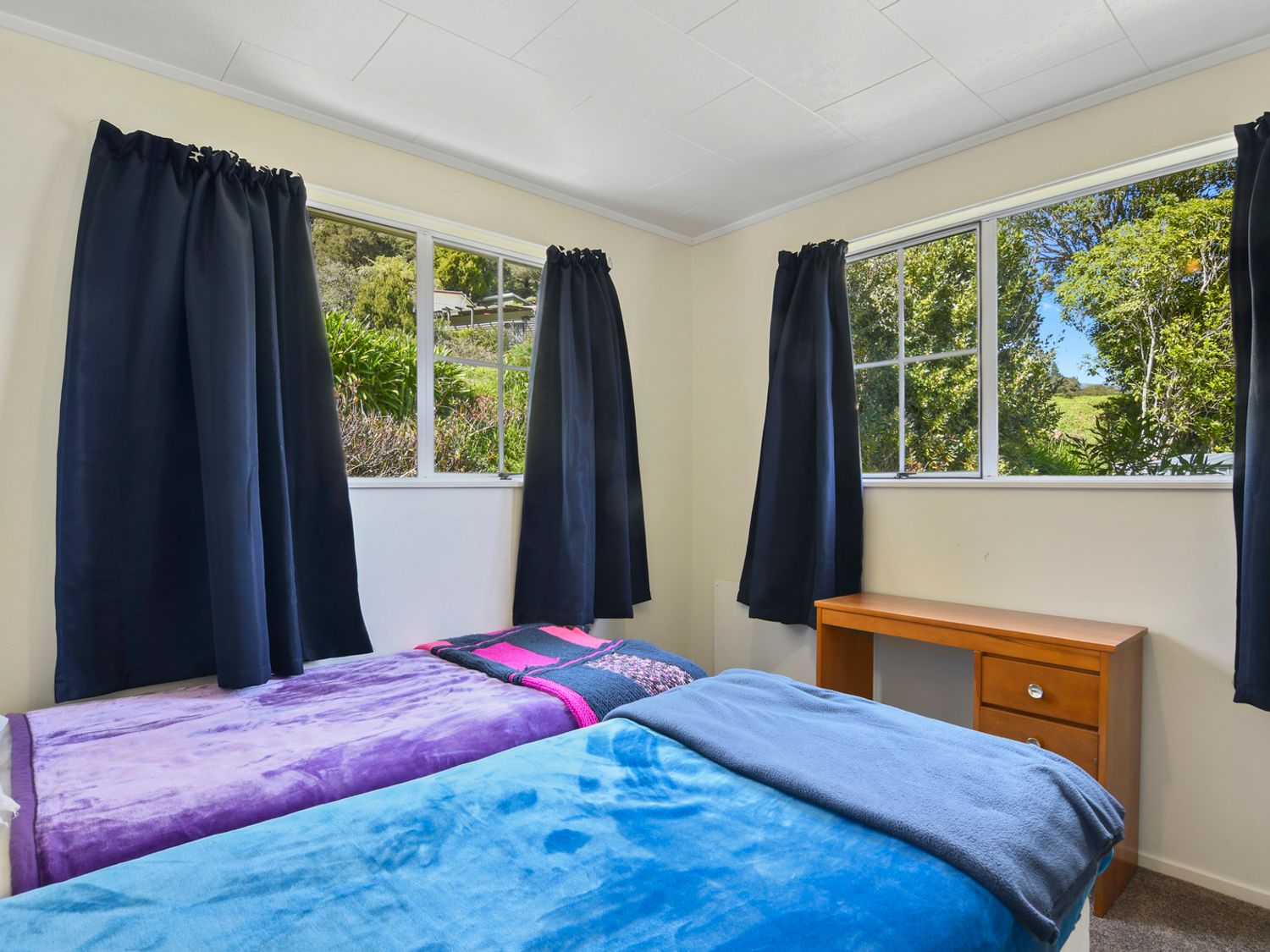 A bedroom with two single beds covered with colorful blankets and a wooden desk under windows with black curtains at Millard House - Marlborough Sounds