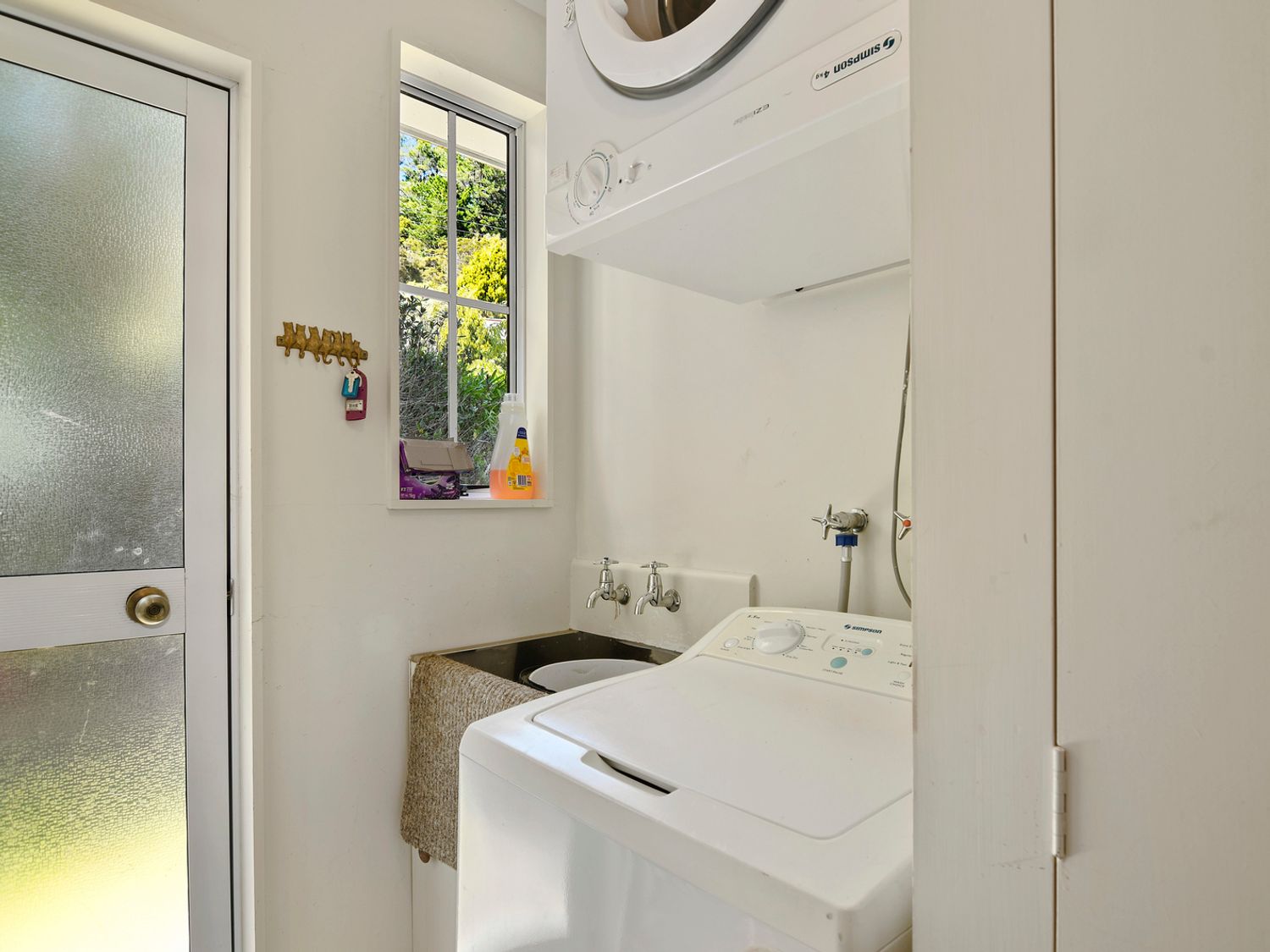 A laundry room with a washing machine, dryer, sink, window, and door at Millard House - Marlborough Sounds