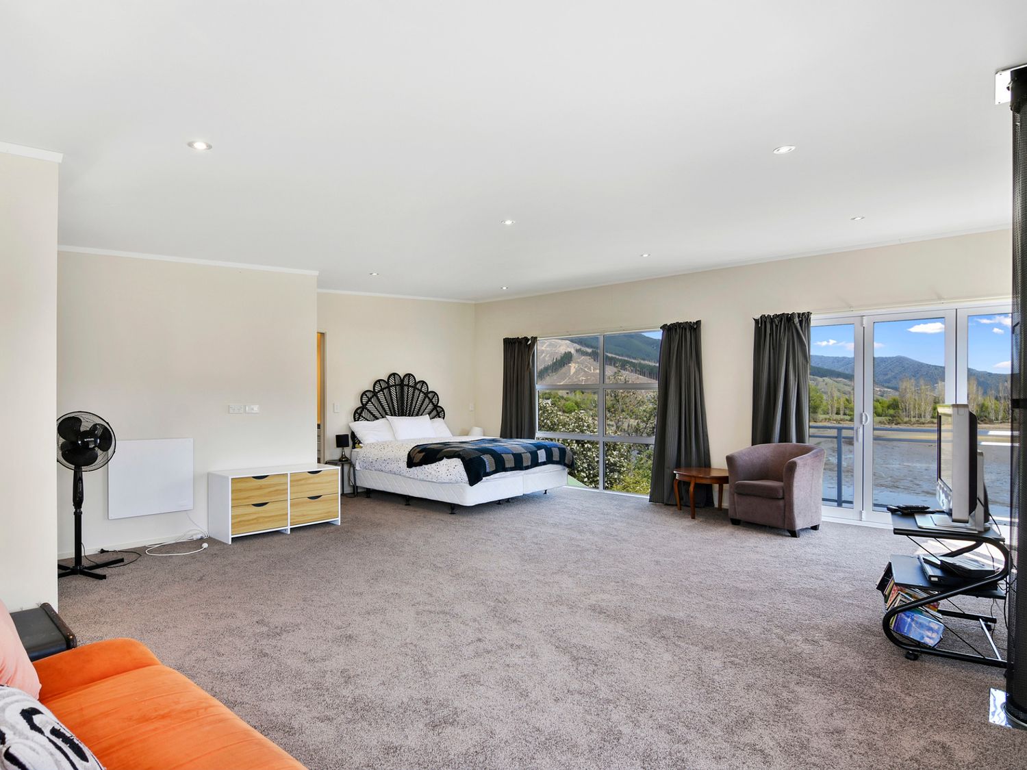 A spacious bedroom with a bed a black fan an orange couch a chair a TV on a stand and large windows with mountain views at Millard House - Marlborough Sounds