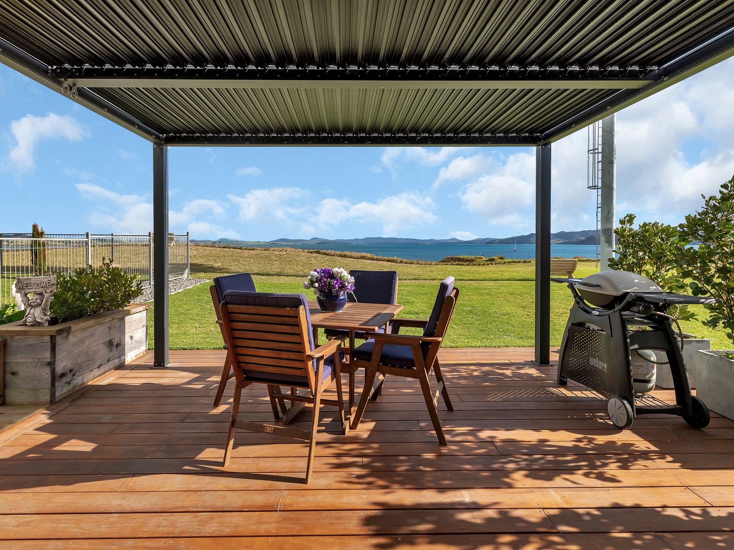 An outdoor patio with wooden table and chairs a barbecue grill and ocean view at The Leading Light - One Tree Point Holiday Home in One Tree Point