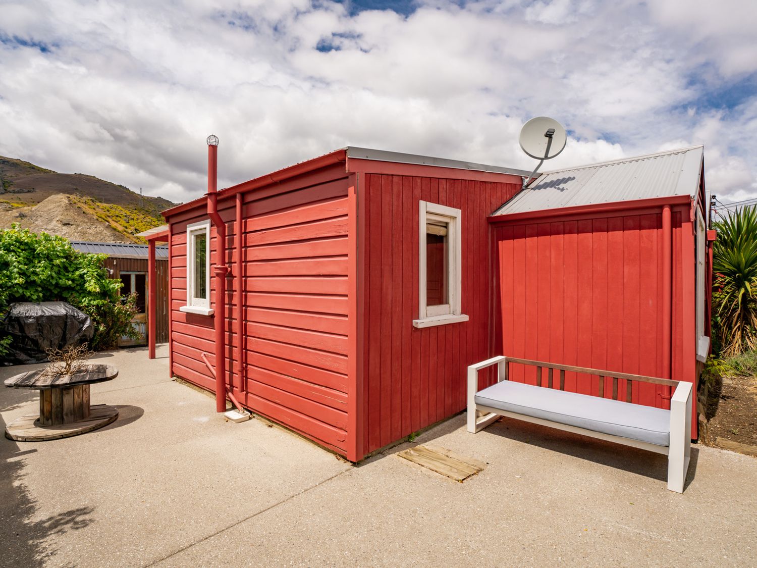 A red wooden house exterior with two windows a gray cushioned bench and a round wooden table on a concrete patio at Cromwell Cutie in Cromwell