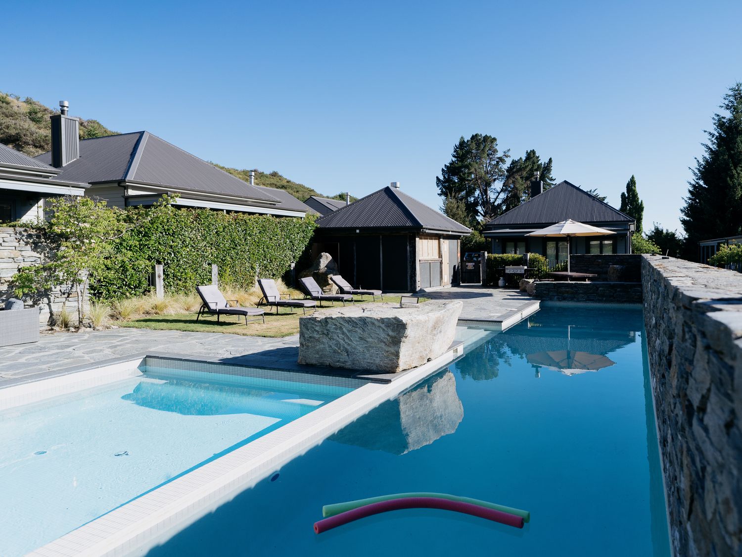 An outdoor pool area with lounge chairs and umbrellas near houses at Cardrona Valley Escape Cardrona Holiday Home