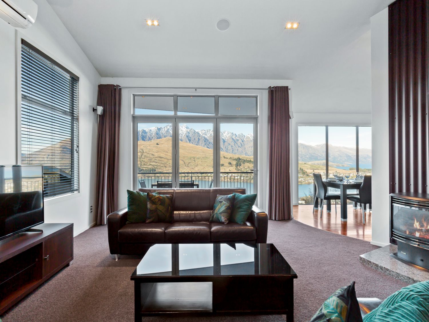 A living room with a leather sofa and coffee table facing large windows showing a mountain view with a dining area and fireplace nearby at Paradise Peaks Queenstown