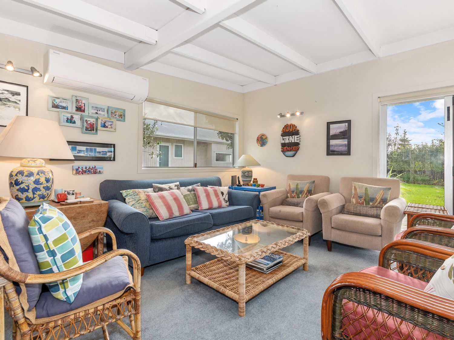 A living room with a blue sofa, beige armchairs, wicker chairs, a glass coffee table, and a lamp at Coastal Cove - Waipu Cove Holiday Home in Waipu Cove