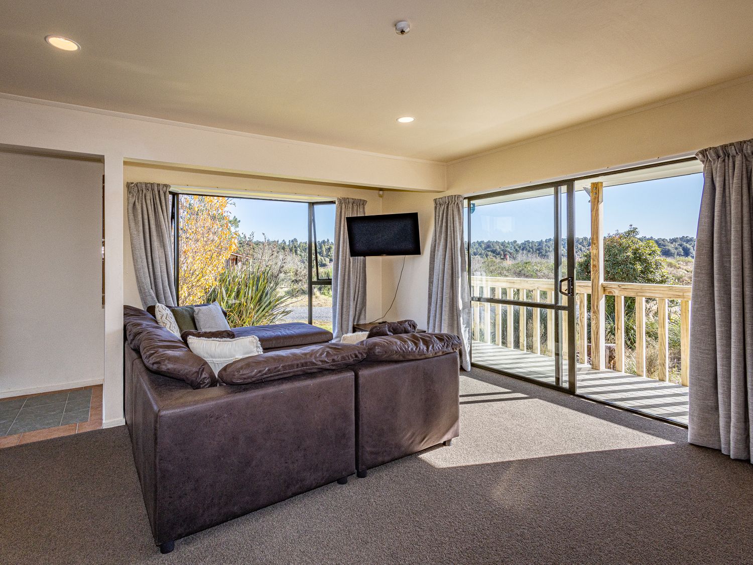 A living room with a brown sectional sofa a mounted television and sliding glass doors opening to a wooden deck at Three Peak Lodge - National Park Holiday Home in National Park