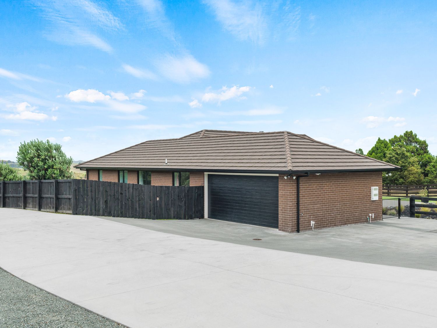 A house with a brown tiled roof and brick walls next to a black wooden fence and a concrete driveway at Mangawhai Maketu Mangawhai
