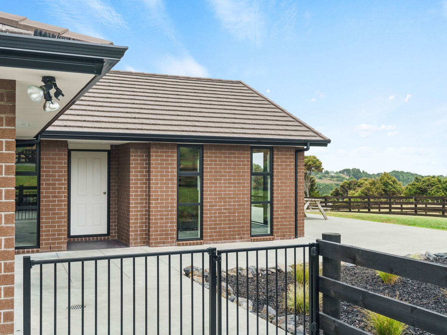 A brick exterior of a house with a white door and black framed windows with a black metal fence and a wooden fence with a picnic table in the background at Mangawhai Maketu Mangawhai